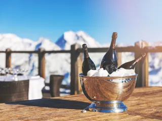 En bøtte med tre champagneflasker utendørs på et trebord med fjell og snø i bakgrunnen