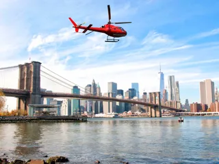 Rødt helikopter flyr over Hudson river med Manhattan og Brooklyn Bridge i bakgrunnen